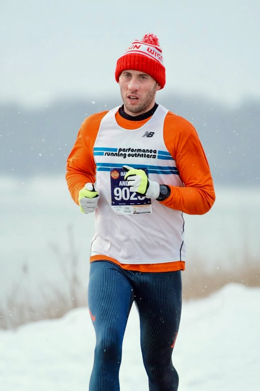 Man running in a snowy landscape wearing a red beanie and orange jacket with a race bib.